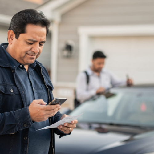 Latin adult reviewing phone by car in Texas driveway, family nearby